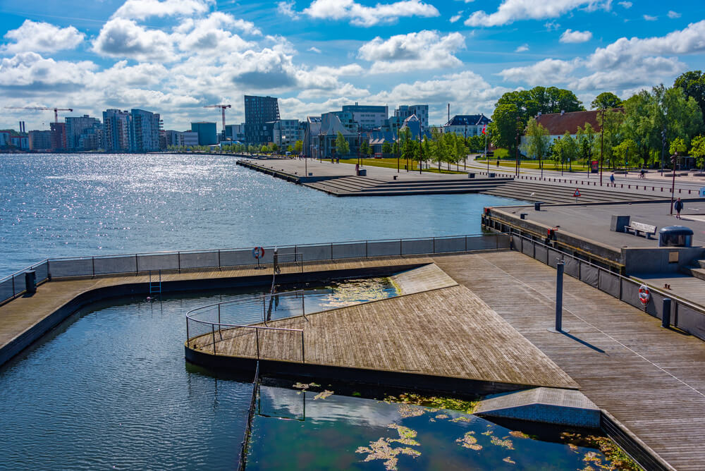 En solrig dag ved havnefronten i Aalborg, Danmark, med en moderne, træbeklædt badebro og et lukket svømmeområde i forgrunden.